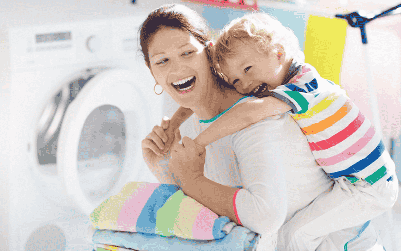 woman and child play in front of a dryer after she folds clean clothes